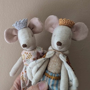 May include: Two plush mouse toys wearing crowns. The mouse on the left is wearing a pink dress with floral print and a silver crown. The mouse on the right is wearing a blue and white striped shirt with a gold crown and a beige cape.