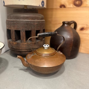 May include: A copper teapot with a black handle and a white and black knob. A dark brown jug with a handle is behind the teapot. A wooden lamp base with a floral patterned lampshade is in the background.