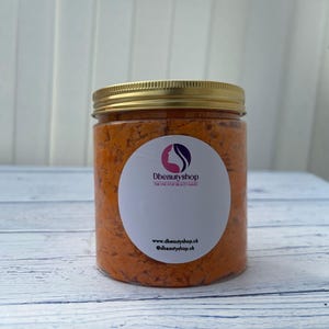 May include: A clear plastic jar with a gold lid filled with an orange product. A white label with a logo and website details is on the jar. The jar is on a light blue wooden surface.