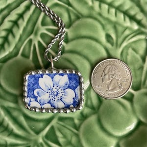 May include: A rectangular pendant featuring a blue and white floral pattern set in a silver-toned beaded frame, attached to a silver-toned rope chain. A United States quarter coin is placed next to the pendant for size comparison.