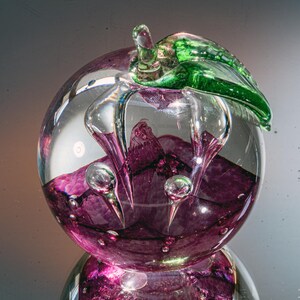 Könnte beinhalten: Ein klarer Glasbriefbeschwerer in Form einer Pflaume, mit einem grünen Blatt und Stiel. Das Innere weist ein tiefviolettes Design mit Luftblasen auf, was ein einzigartiges Dekorationsobjekt ergibt. Die Pflaume spiegelt sich unten.