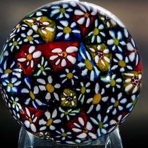 Könnte beinhalten: Ein runder, dekorativer Glas-Briefbeschwerer mit floralem Design. Der Briefbeschwerer hat einen dunkelblauen Hintergrund mit weißen, gelben und roten Blumenmustern. Die Blumen sind in einem mosaikartigen Muster angeordnet.