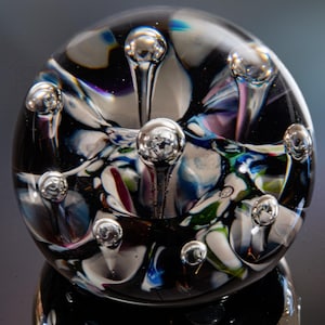 Könnte beinhalten: Ein klarer Glasbriefbeschwerer mit inneren Blasen und farbenfrohen Wirbeln aus Weiß, Blau, Grün und Lila. Der Briefbeschwerer hat eine abgerundete Form mit mehreren kugelförmigen Blasen auf der Oberfläche.