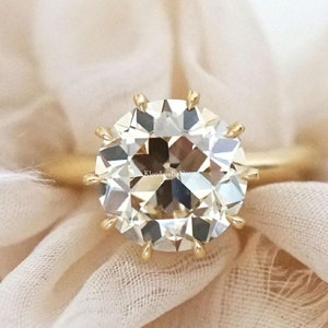 Puede incluir: Un anillo de oro con un gran diamante redondo y claro. El diamante tiene un corte de cojín y está engastado en una banda de oro con un halo de diamantes más pequeños.