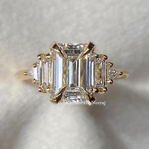 Puede incluir: Un anillo de oro con una gran piedra central de diamante de talla esmeralda y diamantes más pequeños de talla baguette a los lados.