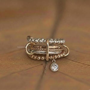 Puede incluir: Un conjunto de anillos apilables con múltiples bandas en tonos plateados, dorados y oro rosa. Los anillos incluyen diseños con cuentas, trenzados y lisos, con un dije de gema transparente colgante. Los anillos se muestran sobre una superficie marrón.