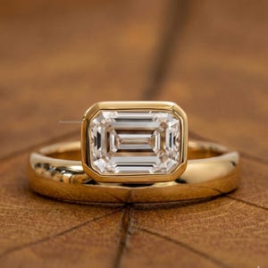 May include: A gold ring with a large, rectangular, clear gemstone. The gemstone is set in a gold bezel. The band of the ring is also gold and has a smooth, rounded design. The ring is on a wooden surface.