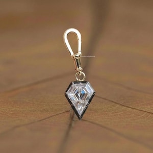 Puede incluir: Un colgante en forma de diamante con un cierre dorado. El colgante presenta una gema clara y facetada engastada en un marco negro. El cierre está unido a la parte superior del colgante.