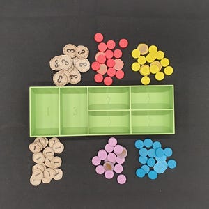 May include: A green plastic organizer with six compartments, filled with various game pieces. There are beige tokens with the number "3" printed on them, red, yellow, pink, and blue circular tokens, and beige tokens with the number "1" printed on them.