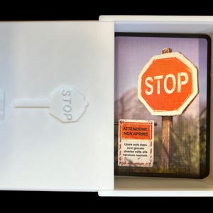 May include: A white box with a red stop sign on the front. The box is open and inside is a smaller box with a picture of a stop sign and a sign that says "Attenzione - Non Aprire" (Attention - Do Not Open). The sign also says "Usare solo dopo aver giocato diverse volte alla versione normale" (Use only after playing several times in the normal version).