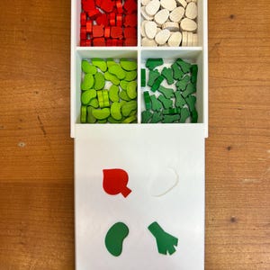 May include: A white plastic tray with four compartments filled with wooden game pieces. The compartments contain red, white, green, and green pieces. The tray also has a smaller compartment with four sample pieces: a red heart, a white circle, a green bean, and a green broccoli.