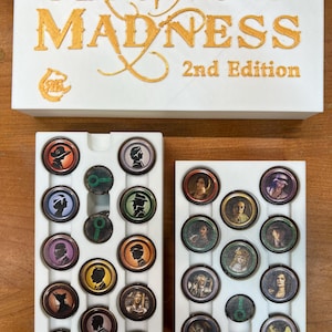 May include: A white game box with gold lettering that reads "Mansions of Madness 2nd Edition." Below the box are two white trays filled with circular game tokens featuring character portraits and symbols. The tokens are in various colors.
