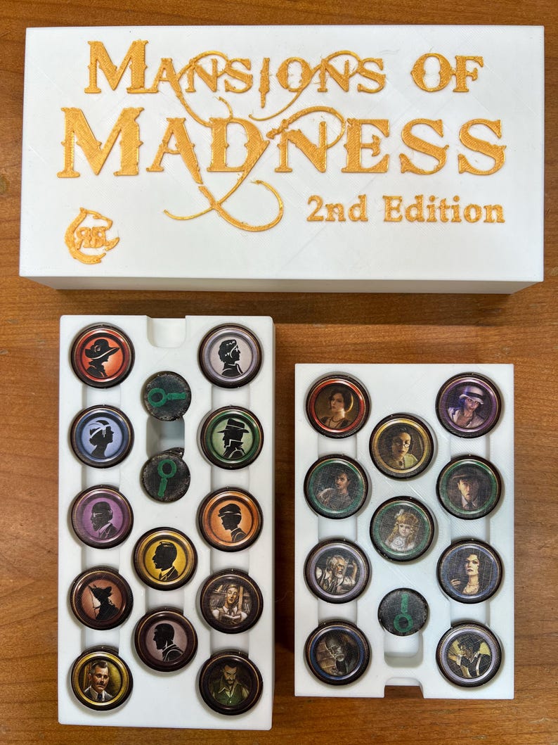 May include: A white game box with gold lettering that reads "Mansions of Madness 2nd Edition." Below the box are two white trays filled with circular game tokens featuring character portraits and symbols. The tokens are in various colors.