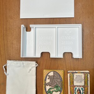 Op de afbeelding: Witte plastic organizer met drie compartimenten voor het opbergen van kaarten. De compartimenten zijn gelabeld met "Victory Region Cards", "Cunning Action Cards" en "Bag". Een witte stoffen tas, een victory region card met een kaart van Devon en een cunning action card met tekst in het Italiaans zijn ook te zien.