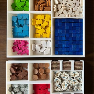 May include: A white organizer with multiple compartments filled with various colored wooden game pieces. The pieces include small houses, trees, and other shapes. There are also some small, round wooden discs and a few small, white tiles with images of tools.