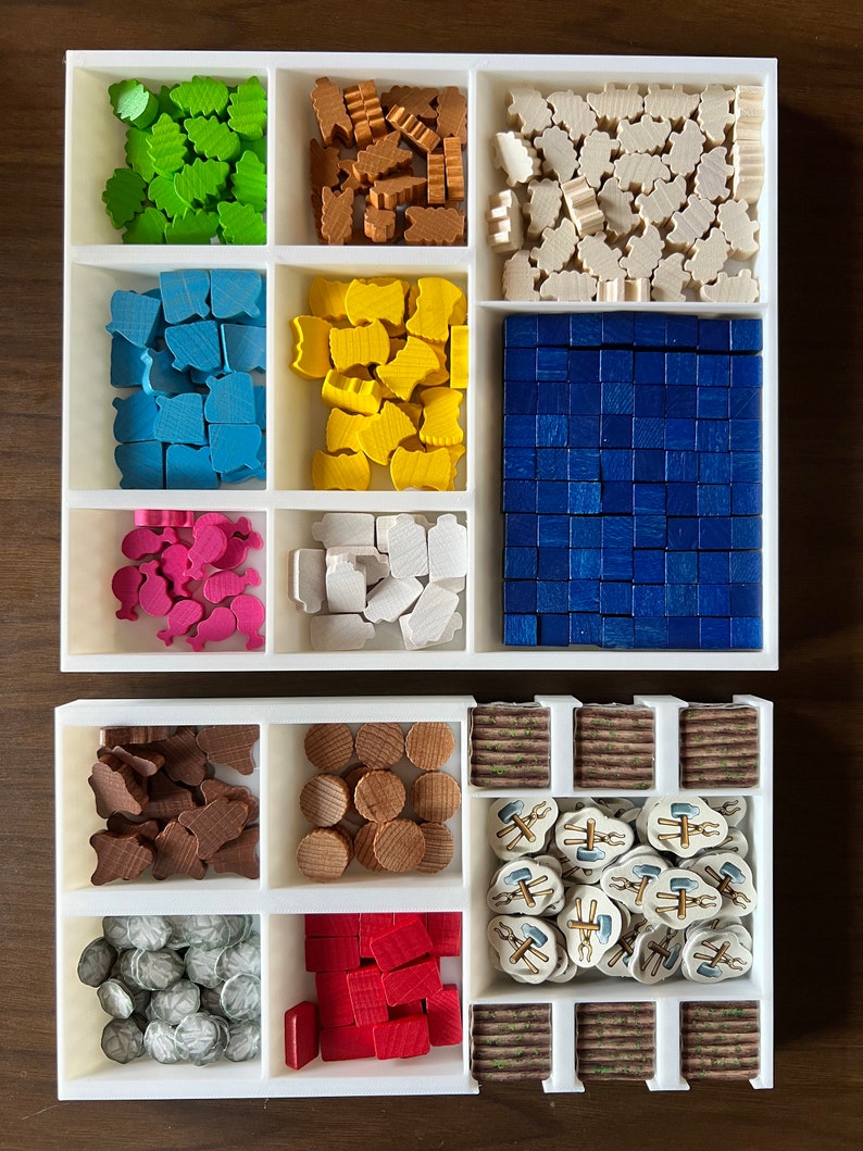 May include: A white organizer with multiple compartments filled with various colored wooden game pieces. The pieces include small houses, trees, and other shapes. There are also some small, round wooden discs and a few small, white tiles with images of tools.