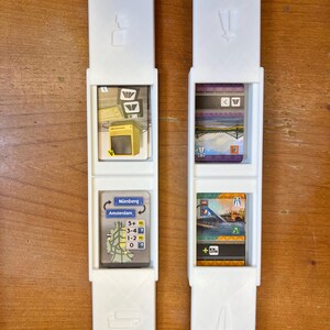 Pode incluir: Dois porta-cart&otilde;es de pl&aacute;stico branco com uma ranhura para um cart&atilde;o no meio. As ranhuras superior e inferior t&ecirc;m um s&iacute;mbolo de um comboio e uma estrada. Os cart&otilde;es nos porta-cart&otilde;es s&atilde;o do jogo de tabuleiro Ticket to Ride. Os cart&otilde;es mostram uma imagem de uma esta&ccedil;&atilde;o de comboios, uma ponte e um mapa de Amesterd&atilde;o e Nuremberga.