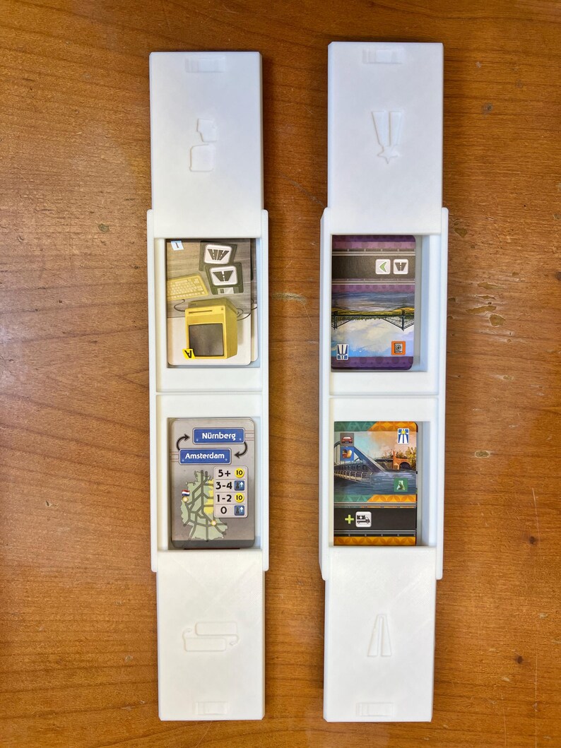 Pode incluir: Dois porta-cart&otilde;es de pl&aacute;stico branco com uma ranhura para um cart&atilde;o no meio. As ranhuras superior e inferior t&ecirc;m um s&iacute;mbolo de um comboio e uma estrada. Os cart&otilde;es nos porta-cart&otilde;es s&atilde;o do jogo de tabuleiro Ticket to Ride. Os cart&otilde;es mostram uma imagem de uma esta&ccedil;&atilde;o de comboios, uma ponte e um mapa de Amesterd&atilde;o e Nuremberga.
