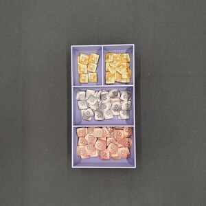 May include: A divided tray filled with game tokens. The top section holds gold-coloured tokens with symbols, the middle section has silver tokens, and the bottom section contains copper-coloured tokens. The tokens are square-shaped.