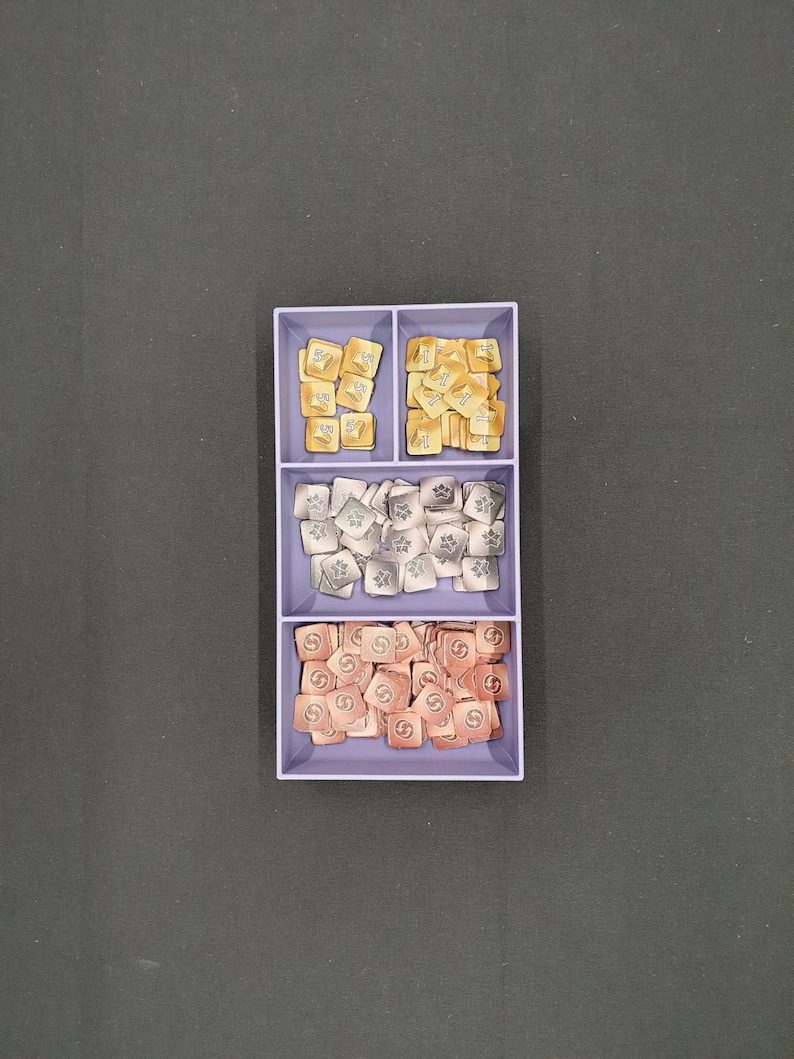 May include: A divided tray filled with game tokens. The top section holds gold-coloured tokens with symbols, the middle section has silver tokens, and the bottom section contains copper-coloured tokens. The tokens are square-shaped.