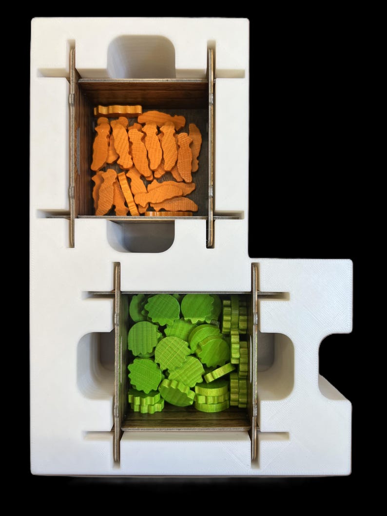 May include: A white foam insert with two compartments for storing game pieces. The top compartment contains orange fish-shaped pieces, and the bottom compartment contains green leaf-shaped pieces.