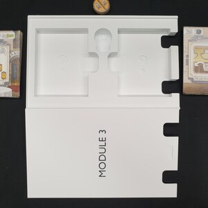 May include: A white game box labeled "MODULE 3" with cutouts for game components. Two stacks of game cards are on either side of the box. A small wooden token sits at the top. The cards feature illustrations and text.