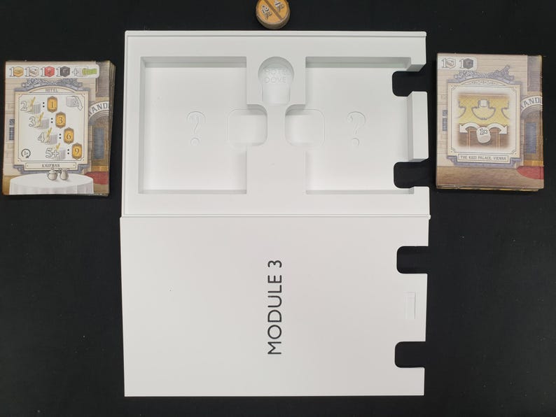 May include: A white game box labeled "MODULE 3" with cutouts for game components. Two stacks of game cards are on either side of the box. A small wooden token sits at the top. The cards feature illustrations and text.