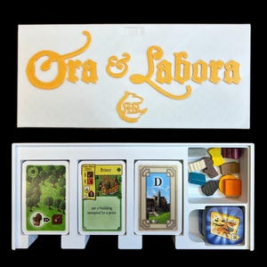 May include: A white box with the words "Ora et Labora" in gold lettering on the top. The box contains a set of cards and wooden pieces for the board game Ora et Labora. The cards are labeled with the names of buildings and the wooden pieces are in different colors and shapes.