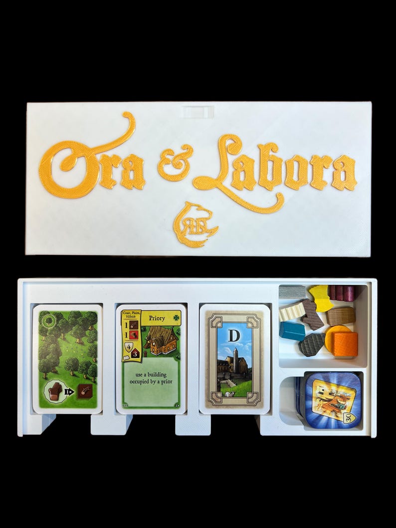 May include: A white box with the words "Ora et Labora" in gold lettering on the top. The box contains a set of cards and wooden pieces for the board game Ora et Labora. The cards are labeled with the names of buildings and the wooden pieces are in different colors and shapes.