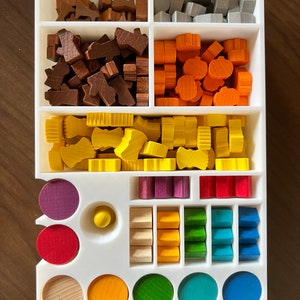 May include: A white organizer with compartments for storing various wooden game pieces. The pieces are in different colors, including brown, gray, orange, yellow, purple, red, green, blue, and natural wood. There are also round wooden pieces in different colors.