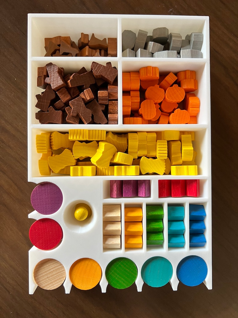 May include: A white organizer with compartments for storing various wooden game pieces. The pieces are in different colors, including brown, gray, orange, yellow, purple, red, green, blue, and natural wood. There are also round wooden pieces in different colors.