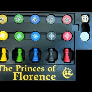 Op de afbeelding: Een zwart spelbord met de titel "The Princes of Florence" en een logo. Het bord heeft vakjes voor spelstukken, waaronder vijf kleine gele, groene, blauwe, zwarte en rode stukken. Er zijn ook vijf vakjes voor tokens met het nummer 50 erop en vijf vakjes voor tokens met een stervormig ontwerp.