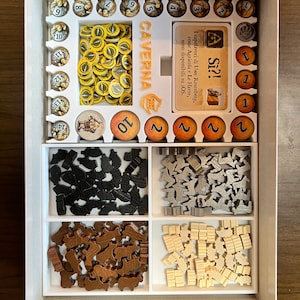 May include: A white foam insert with compartments for storing game pieces for the board game Caverna: The Cave Farmers. The insert includes compartments for wooden meeples, wooden cubes, and plastic tokens. The insert also includes a card holder for the game's rulebook.