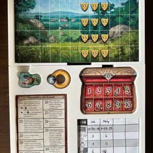 May include: A game board with a colorful illustration of a countryside scene. The board includes a wooden chest with numbered compartments, a score sheet with the names "Linh" and "Micky", and a set of rules in Italian. The game appears to be a strategy board game.