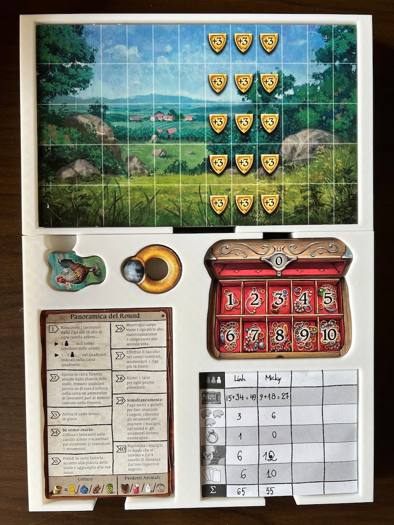 May include: A game board with a colorful illustration of a countryside scene. The board includes a wooden chest with numbered compartments, a score sheet with the names "Linh" and "Micky", and a set of rules in Italian. The game appears to be a strategy board game.