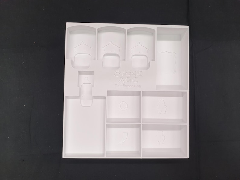 Puede incluir: Un organizador de juego de pl&aacute;stico blanco para el juego de mesa Stone Age: The Expansion. El organizador tiene varios compartimentos de diferentes formas y tama&ntilde;os, incluyendo espacios para piezas y cartas del juego. El texto "Stone Age: The Expansion" es visible.