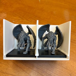 May include: Two gray miniature figures of a creature with wings, tentacles, and a humanoid body. The figures are displayed in a white and black display case, set against a wooden surface. The figures are detailed and appear to be from a fantasy or horror genre.