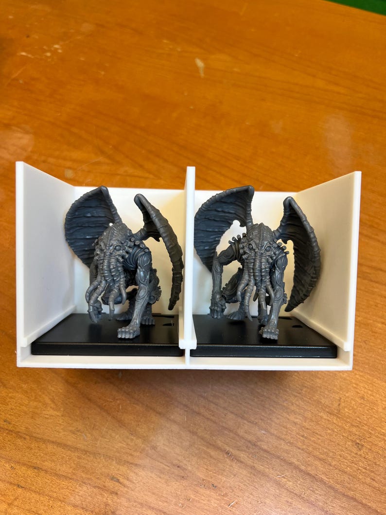 May include: Two gray miniature figures of a creature with wings, tentacles, and a humanoid body. The figures are displayed in a white and black display case, set against a wooden surface. The figures are detailed and appear to be from a fantasy or horror genre.