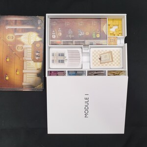 May include: An overhead shot of a board game box and components. The box is white with "MODULE I" printed on the front. Inside are various game pieces, cards, and a game board with a detailed illustration.