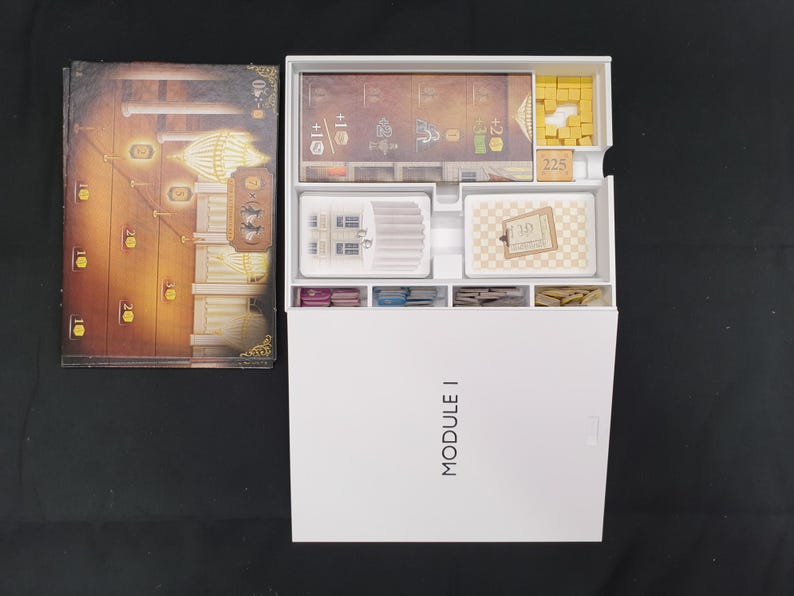 May include: An overhead shot of a board game box and components. The box is white with "MODULE I" printed on the front. Inside are various game pieces, cards, and a game board with a detailed illustration.