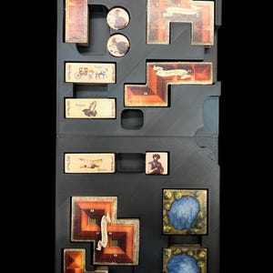 May include: A black storage box with compartments for a board game. The box contains various game pieces, including a small wooden carriage, a blue and green lake tile, and a brown tile with the word "Universita" on it.