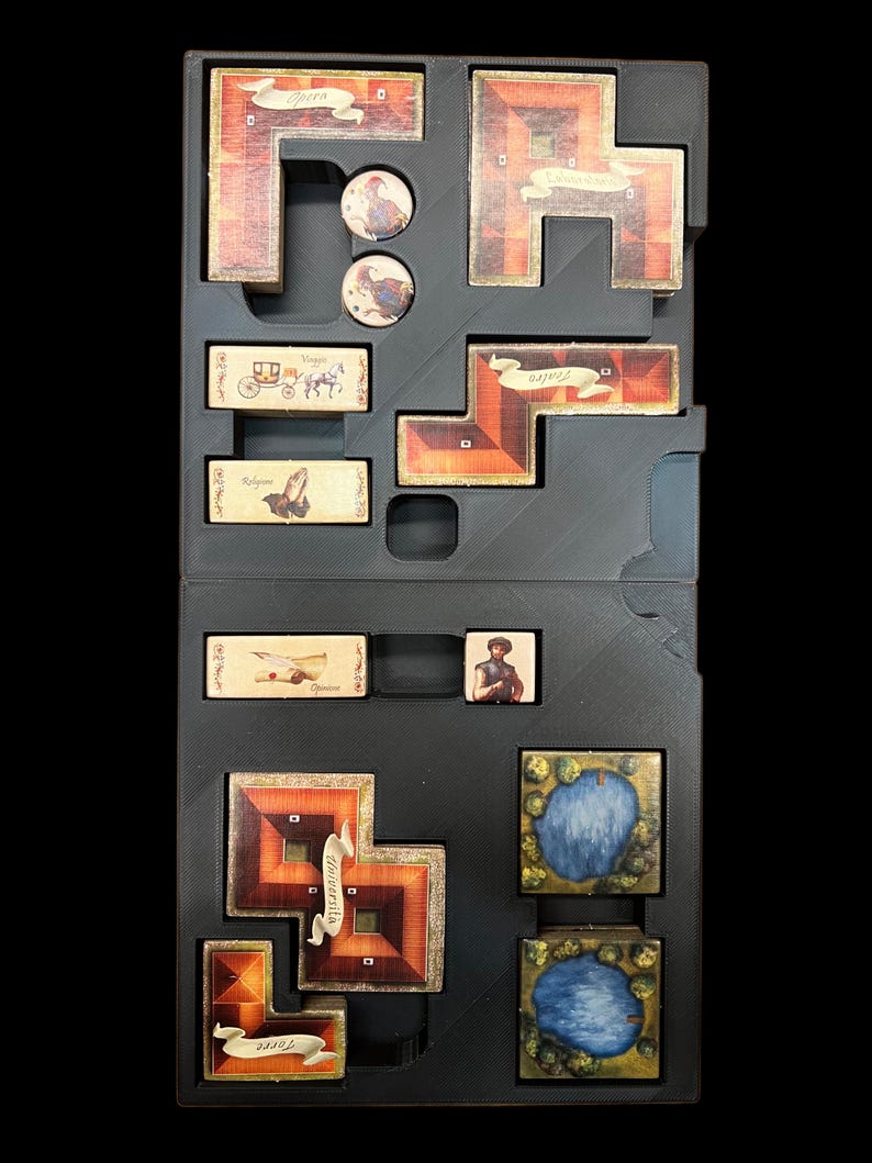 May include: A black storage box with compartments for a board game. The box contains various game pieces, including a small wooden carriage, a blue and green lake tile, and a brown tile with the word "Universita" on it.