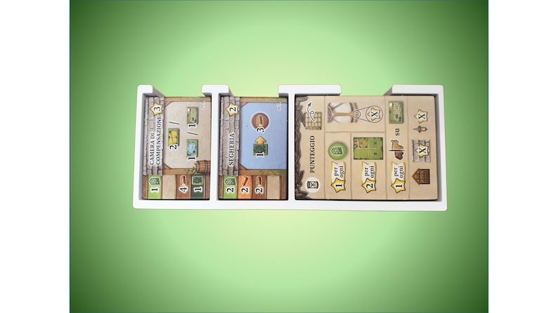 May include: A white organizer with five compartments for storing game cards. The cards are labeled "Camera di Compensazione", "Segheria", "Punteggio", and have numbers and symbols on them.