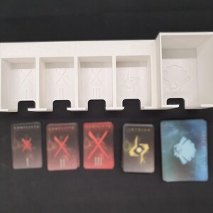 May include: A white plastic game organiser with five compartments and a larger section. Below are five game cards with red, yellow, and blue designs. The cards have Roman numerals and symbols.