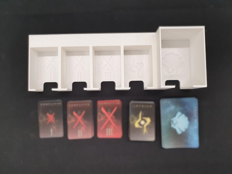 May include: A white plastic game organiser with five compartments and a larger section. Below are five game cards with red, yellow, and blue designs. The cards have Roman numerals and symbols.