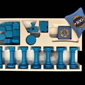 May include: A white foam insert with blue plastic game pieces. The insert has compartments for storing different shapes and sizes of pieces. There are also seven blue columns. A blue dice with a +1 symbol and a blue pillow with the text +100 are also included.