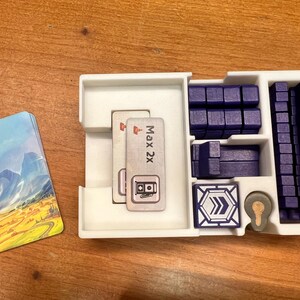 Pode incluir: Um organizador de pl&aacute;stico branco com compartimentos para guardar pe&ccedil;as de jogo. O organizador tem dois cart&otilde;es com o texto "Max 2x" e um s&iacute;mbolo de um posto de gasolina. Tamb&eacute;m h&aacute; blocos de pl&aacute;stico azul e uma placa de pl&aacute;stico azul com um s&iacute;mbolo de seta.