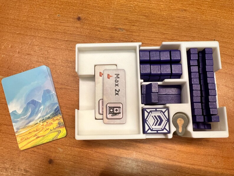 Pode incluir: Um organizador de pl&aacute;stico branco com compartimentos para guardar pe&ccedil;as de jogo. O organizador tem dois cart&otilde;es com o texto "Max 2x" e um s&iacute;mbolo de um posto de gasolina. Tamb&eacute;m h&aacute; blocos de pl&aacute;stico azul e uma placa de pl&aacute;stico azul com um s&iacute;mbolo de seta.