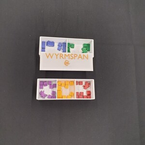 May include: Two white rectangular game component organizers with coloured game pieces. The top organizer has blue, green, and purple pieces. The bottom organizer has purple, yellow, and red pieces. The game name "WYRMSPAN" is printed on the top organizer.