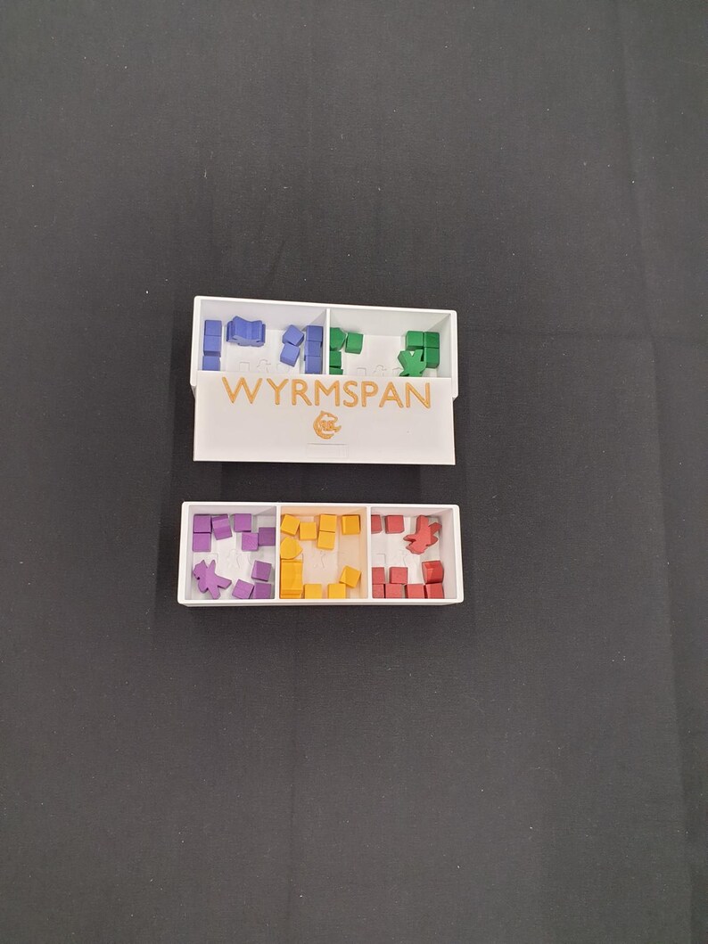 May include: Two white rectangular game component organizers with coloured game pieces. The top organizer has blue, green, and purple pieces. The bottom organizer has purple, yellow, and red pieces. The game name "WYRMSPAN" is printed on the top organizer.